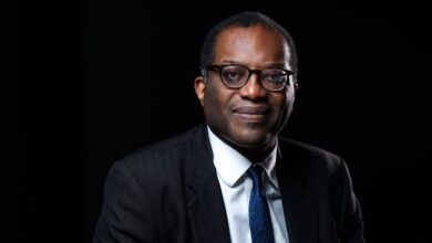 Kwasi Kwarteng wearing a dark suit and glasses, posed against a blue background with the text ‘Kwasi Kwarteng Net Worth’ displayed beside him.”