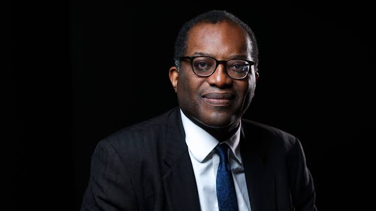 Kwasi Kwarteng wearing a dark suit and glasses, posed against a blue background with the text ‘Kwasi Kwarteng Net Worth’ displayed beside him.”